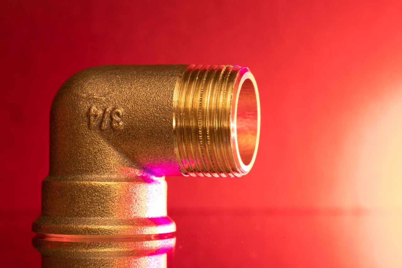 Close-up of a shiny brass pipe elbow fitting against a vibrant red backdrop, showcasing industrial elegance.